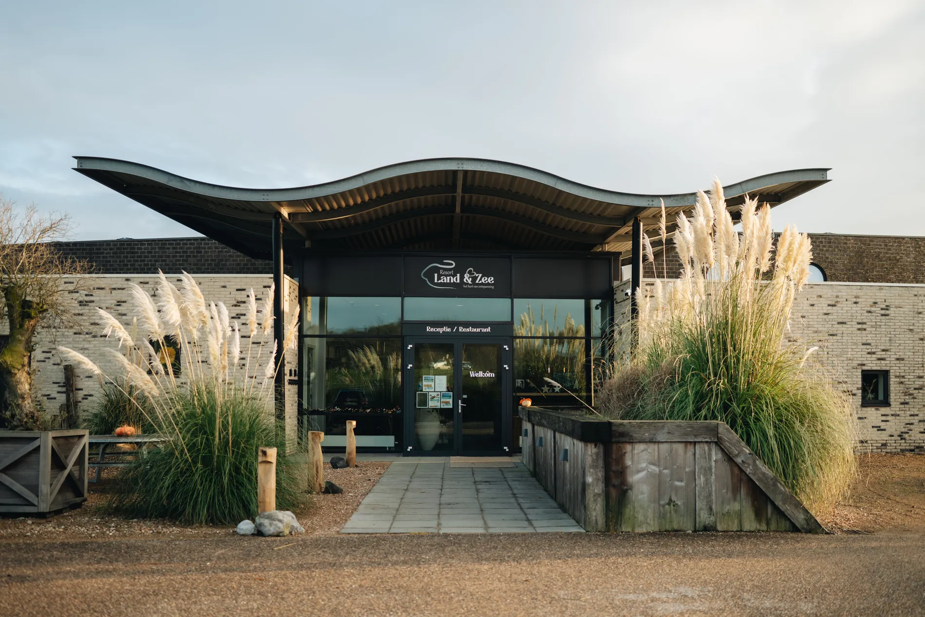 De hoofdingang van de receptie en het restaurant van Resort Land & Zee, gekenmerkt door modern design, pampasgras en het herkenbare golvende dak.